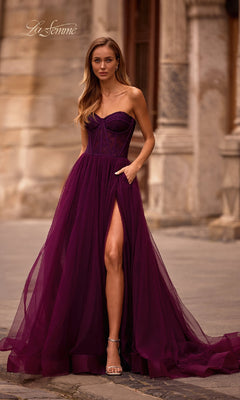 A woman wearing the La Femme 32579 Strapless Long Prom Ball Gown, featuring a floral corset-style bodice and thigh-high slit, stands outside on stone pavement in front of a historic building.