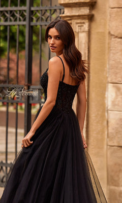 A woman with long brown hair in a sleeveless Beaded-Bodice Long Prom Ball Gown: La Femme 32594 stands outdoors by a wrought iron gate and stone wall, looking over her shoulder.