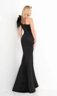A woman in the Jovani 32602 One-Shoulder Mermaid Prom Gown, featuring a fitted black silhouette and large bow detail, stands against a white wall with her back to the camera.