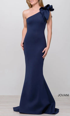 A woman models the Jovani 32602 One-Shoulder Mermaid Prom Gown in navy blue, featuring a dramatic shoulder bow; “Jovani” is displayed in the bottom right corner.