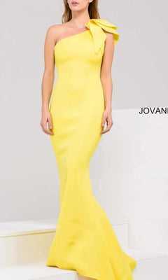 A woman stands elegantly on white steps in the Jovani 32602 One-Shoulder Mermaid Prom Gown, a fitted, floor-length yellow dress featuring a large bow on the shoulder.