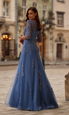 A woman stands outdoors in the La Femme 32612, a long blue beaded formal dress with sheer sleeves and floral embroidery—an elegant choice for a mother of the bride, set against a historic city square backdrop.