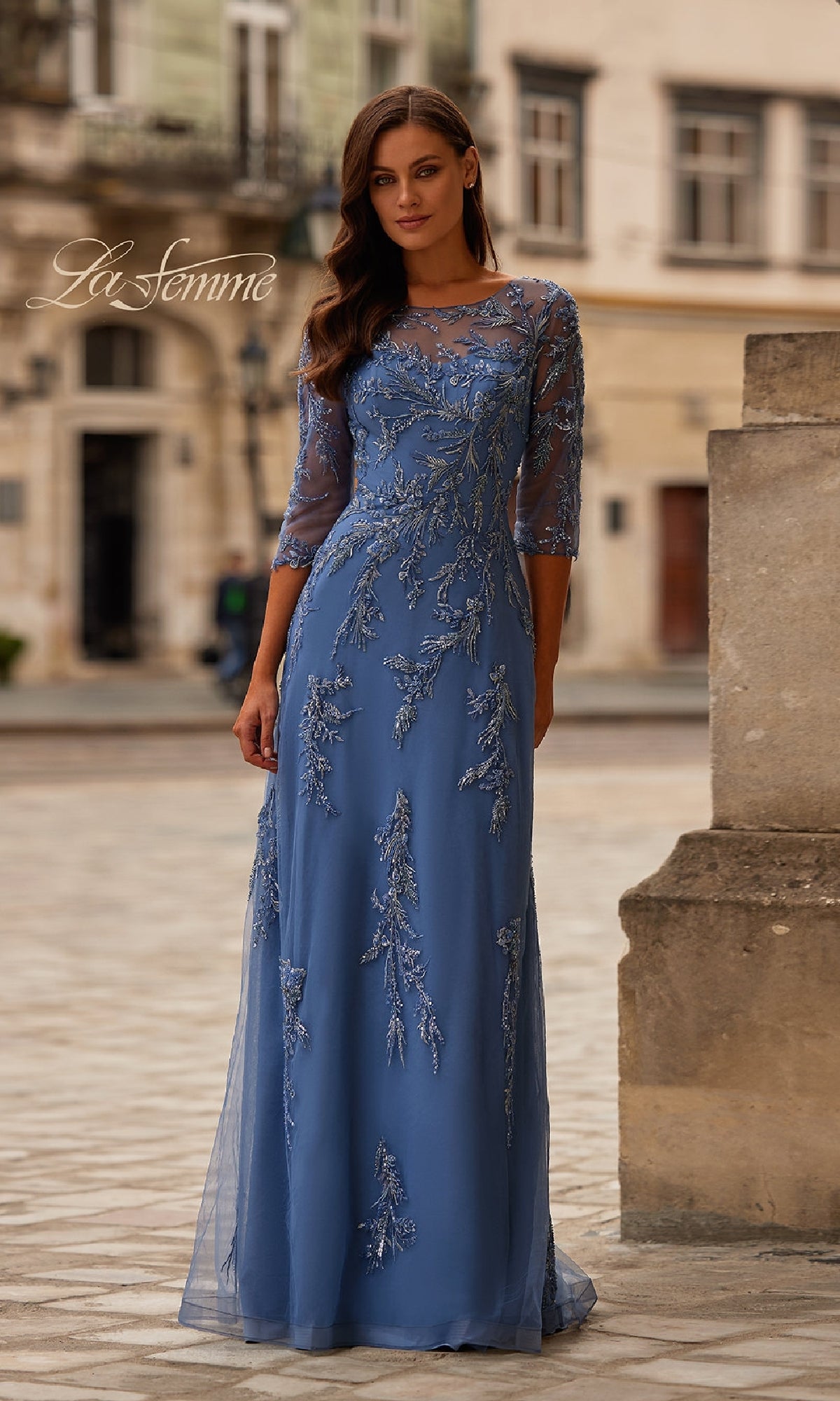A woman stands outdoors in the La Femme 32612—an elegant long blue beaded formal dress with sheer sleeves and floral embroidery, set against historic buildings. A stunning choice for a mother of the bride look.