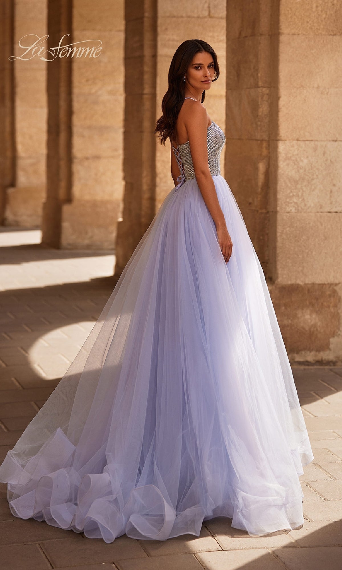 A woman poses near stone arches outdoors in the La Femme 32633 Beaded-Corset Periwinkle Prom Gown, featuring a rhinestone-beaded mesh bodice and a full tulle skirt.