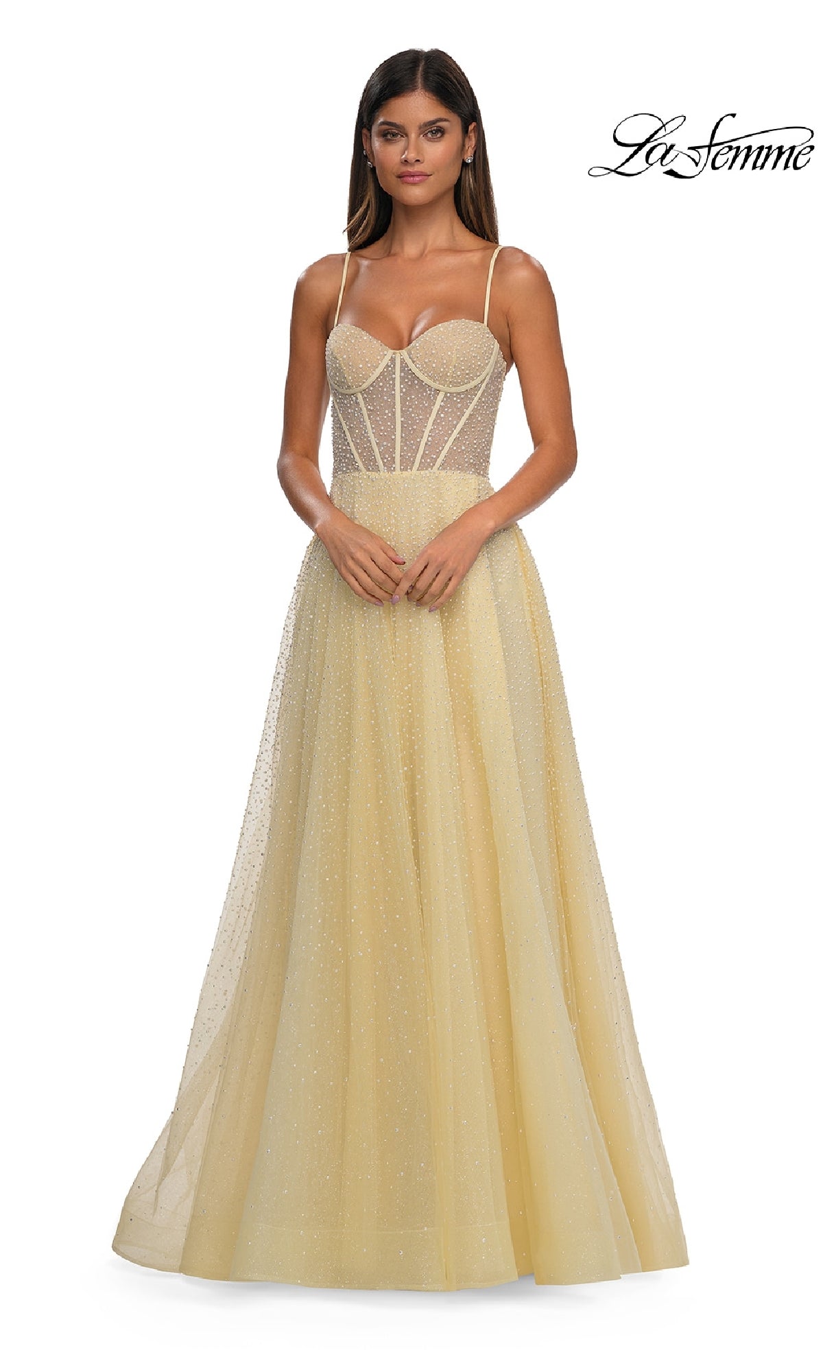 A woman in the Pearl Classic Long Prom Dress: La Femme 32644—a light yellow, floor-length a-line gown with sheer beaded bodice and thin straps—stands with hands clasped. The 