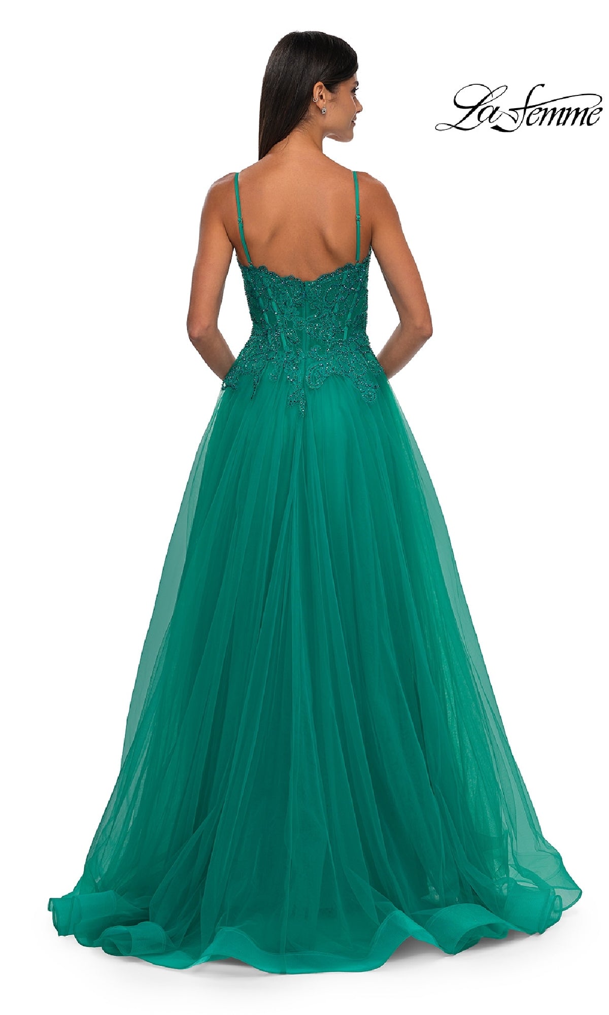 A woman in a floor-length emerald green La Femme 32646 prom ball gown with pockets, thin straps, and an embellished bodice stands with her back to the camera.