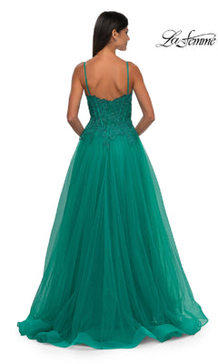 A woman in a floor-length emerald green La Femme 32646 prom ball gown with pockets, thin straps, and an embellished bodice stands with her back to the camera.
