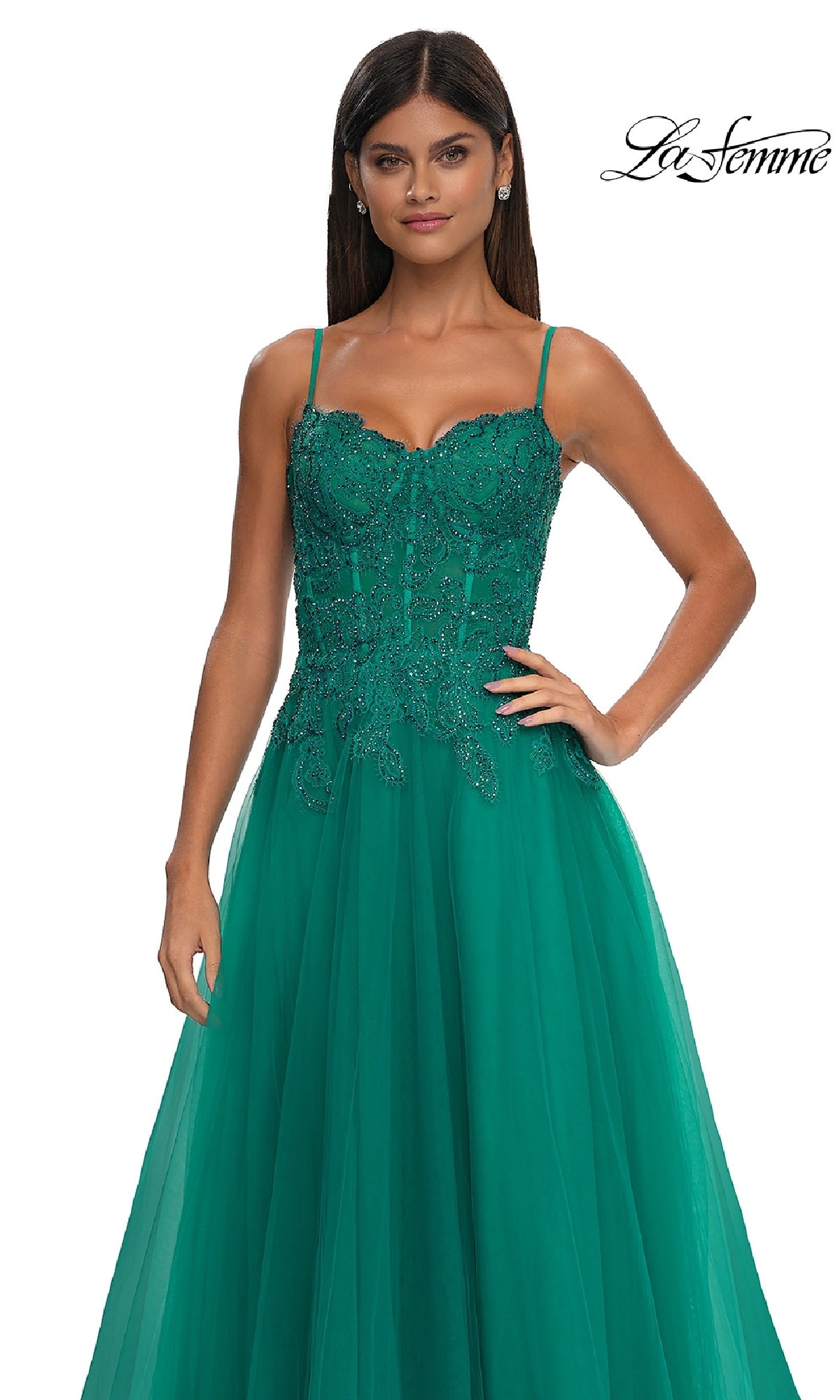 A woman poses with one hand on her hip in a sleeveless emerald green floral beaded gown with sheer overlay and pockets (La Femme 32646), against a white background. 