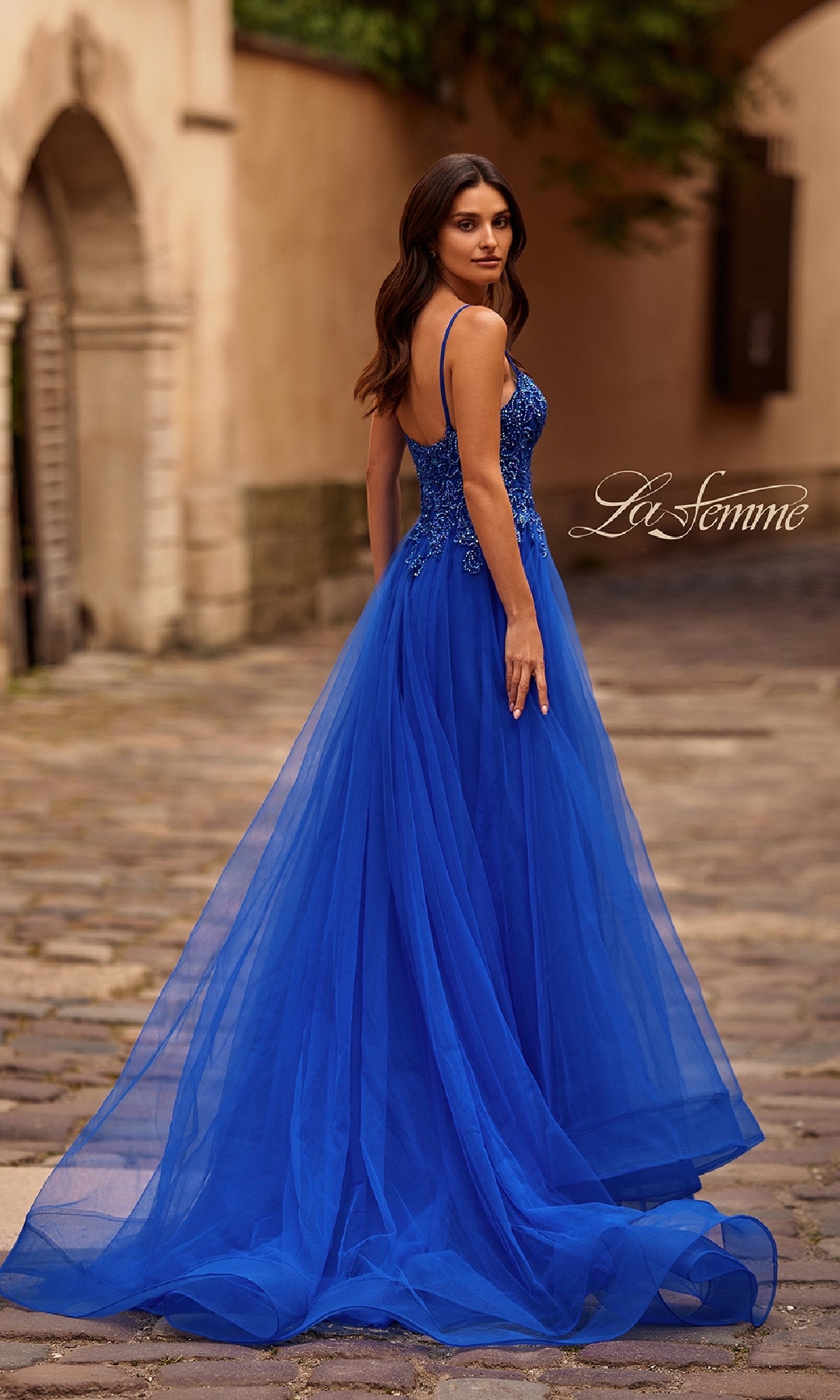 A woman stands on a cobblestone street, looking over her shoulder in the La Femme 32646 long prom ball gown with pockets and a tulle skirt, elegant in royal blue.