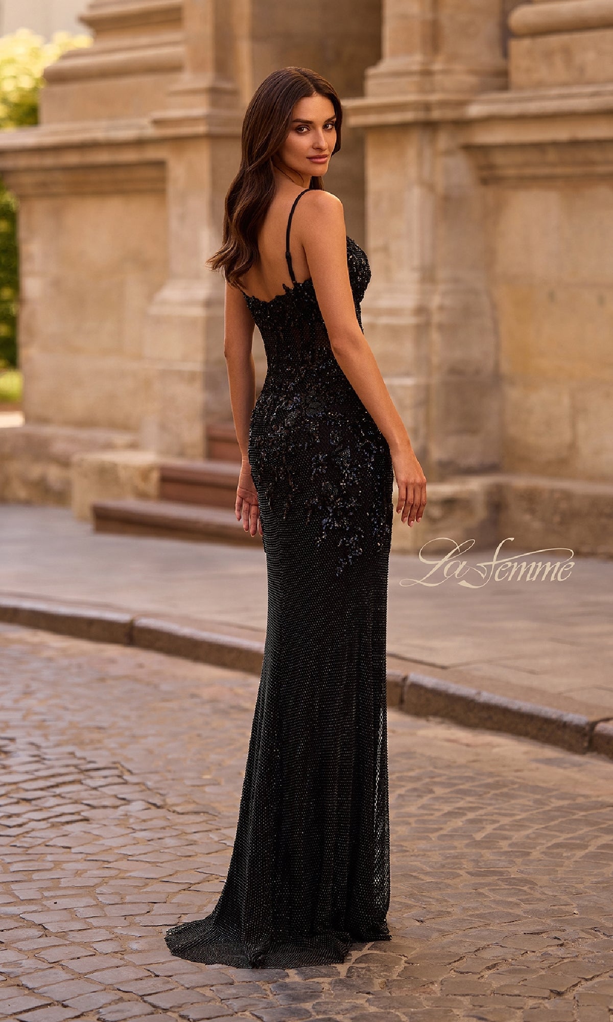 A woman poses outdoors on a cobblestone street in the La Femme 32648, a long black prom dress with a sheer corset bodice, framed by historical architecture in the background.