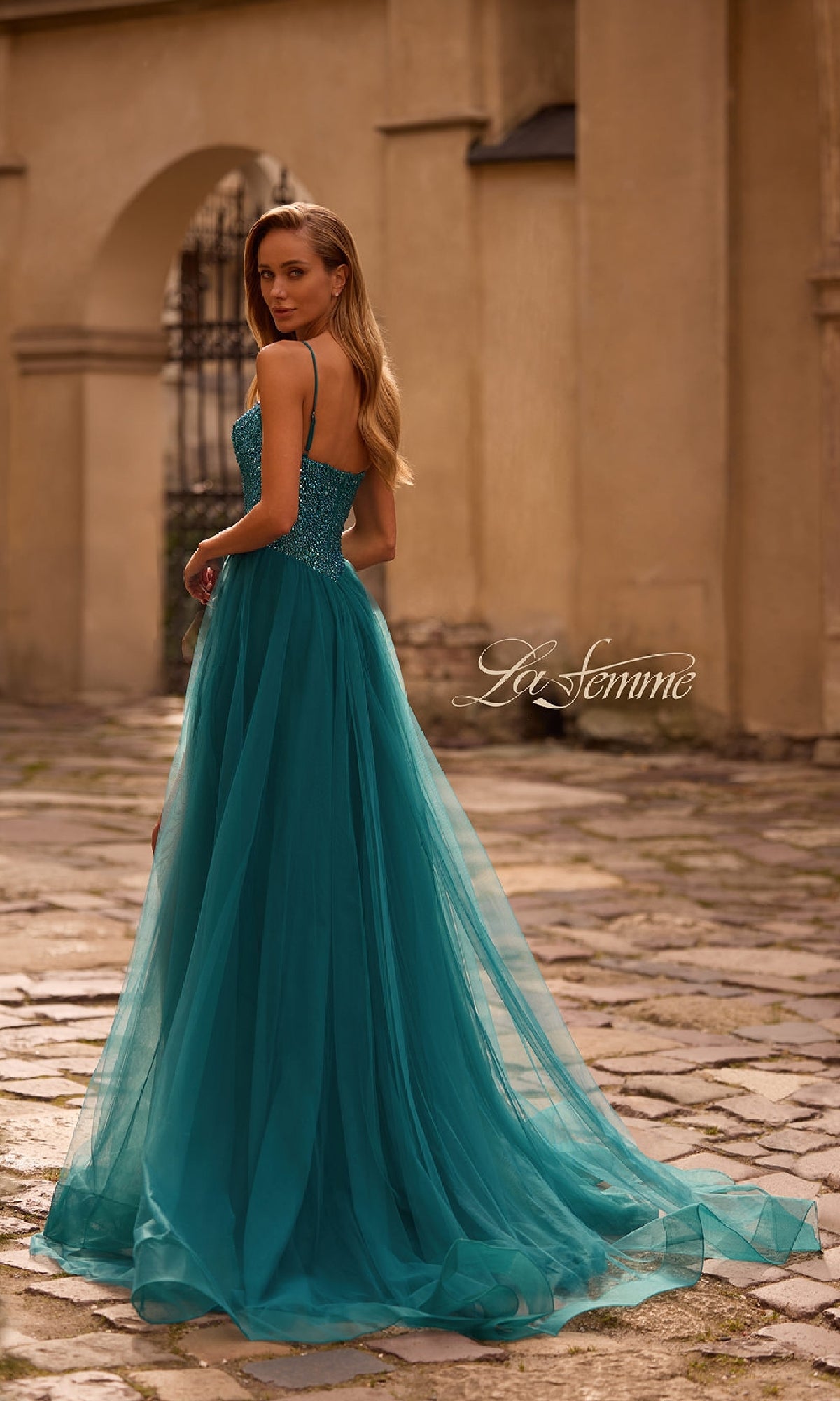 A woman stands on a cobblestone path in the La Femme 32668 Beaded-Bodice Long Prom Ball Gown, featuring a teal beaded bodice and flowing tulle skirt, glancing over her shoulder.