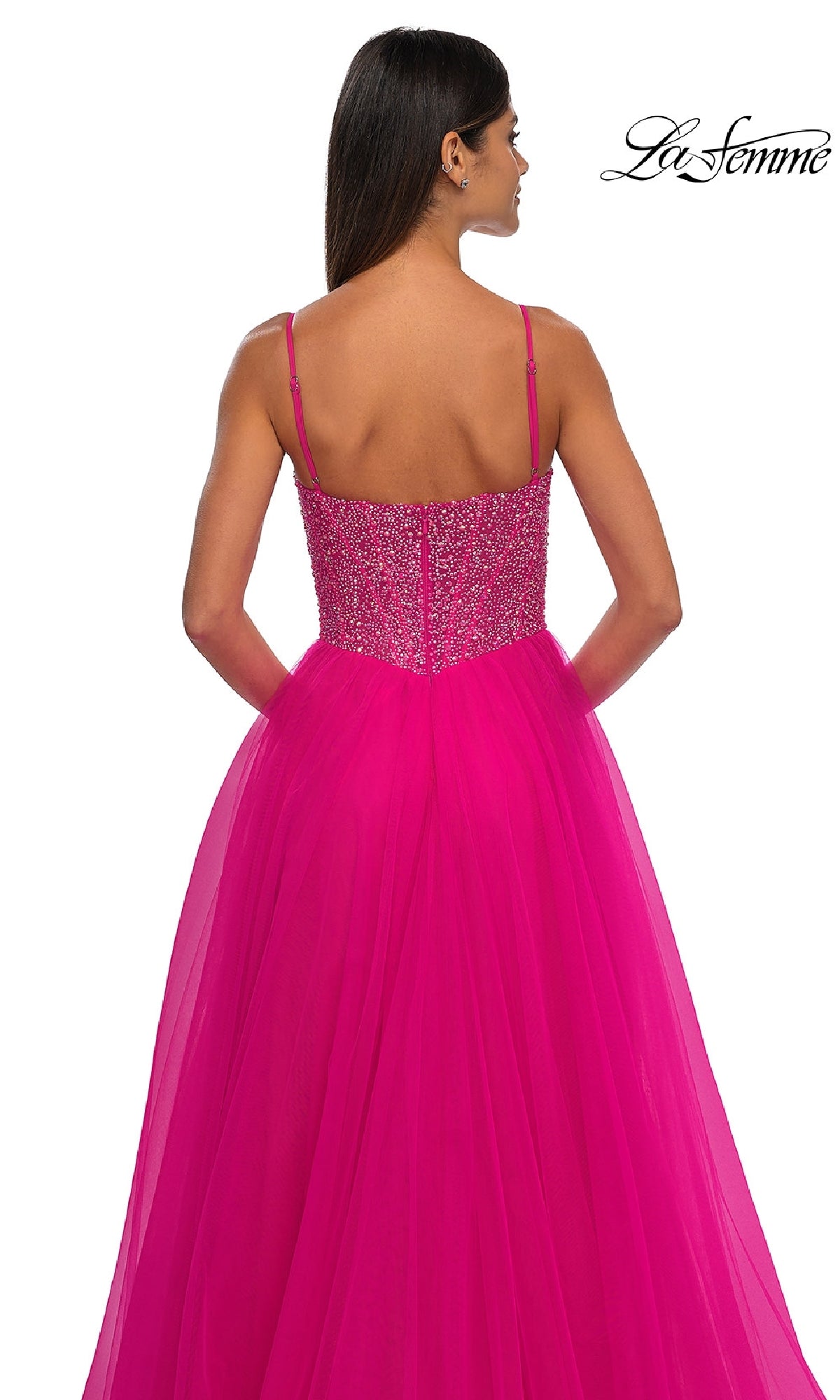 Woman in a bright pink La Femme 32668 prom ball gown with a beaded v-neck bodice and thin straps, shown from the back on a white background. 
