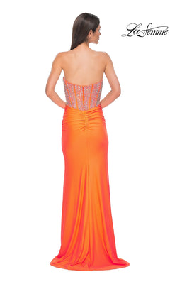 A woman stands facing away, wearing the La Femme 32669: a vibrant orange strapless long corset prom dress with ruched details, sheer beaded corset back, and "La Femme" logo in the top right corner.