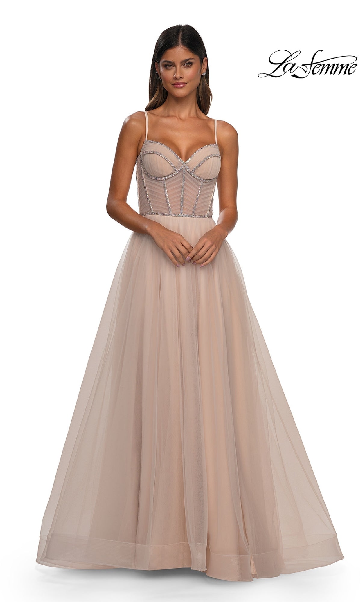 A woman wears the Long Corset Prom Ball Gown: La Femme 32675, a light beige, floor-length A-line dress with thin straps and a structured bodice, standing against a plain white background. 