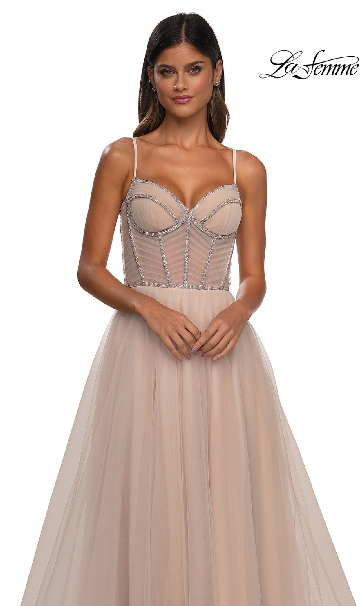 A woman wears the Long Corset Prom Ball Gown: La Femme 32675 in light beige, featuring a sleeveless a-line design with sheer bodice and thin straps, standing against a white background. 