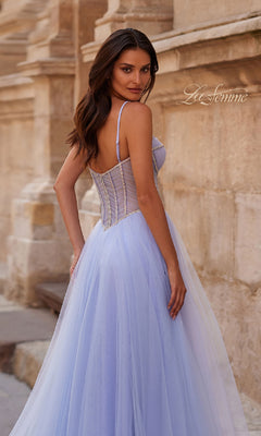 A woman stands outdoors in a light blue, sleeveless Long Corset Prom Ball Gown: La Femme 32675, looking over her shoulder with a stone building behind her. "La Femme" is visible on the dress.