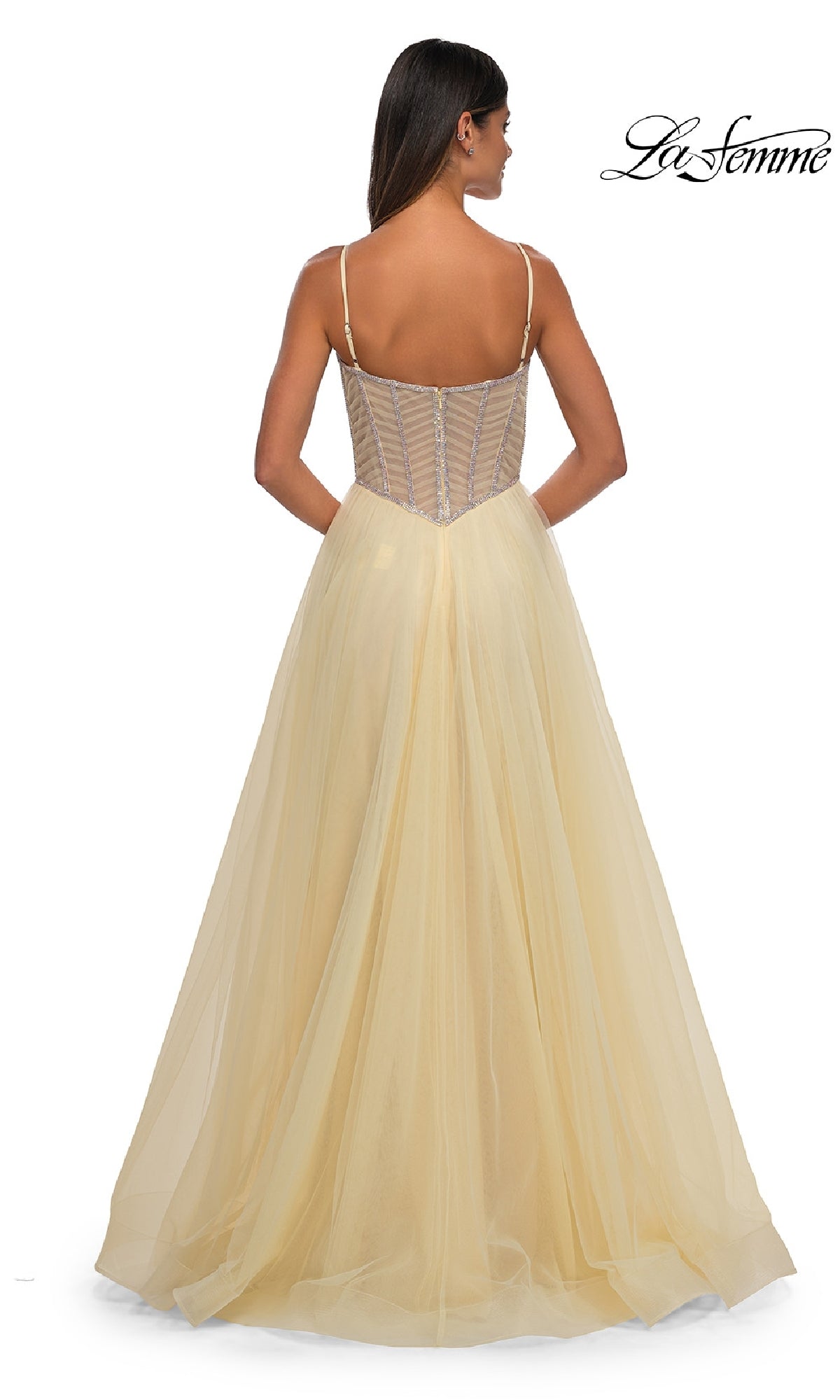 A woman is wearing the Long Corset Prom Ball Gown: La Femme 32675, a light yellow, floor-length dress with a sheer fitted bodice and spaghetti straps, facing away from the camera. This elegant gown exudes effortless sophistication.