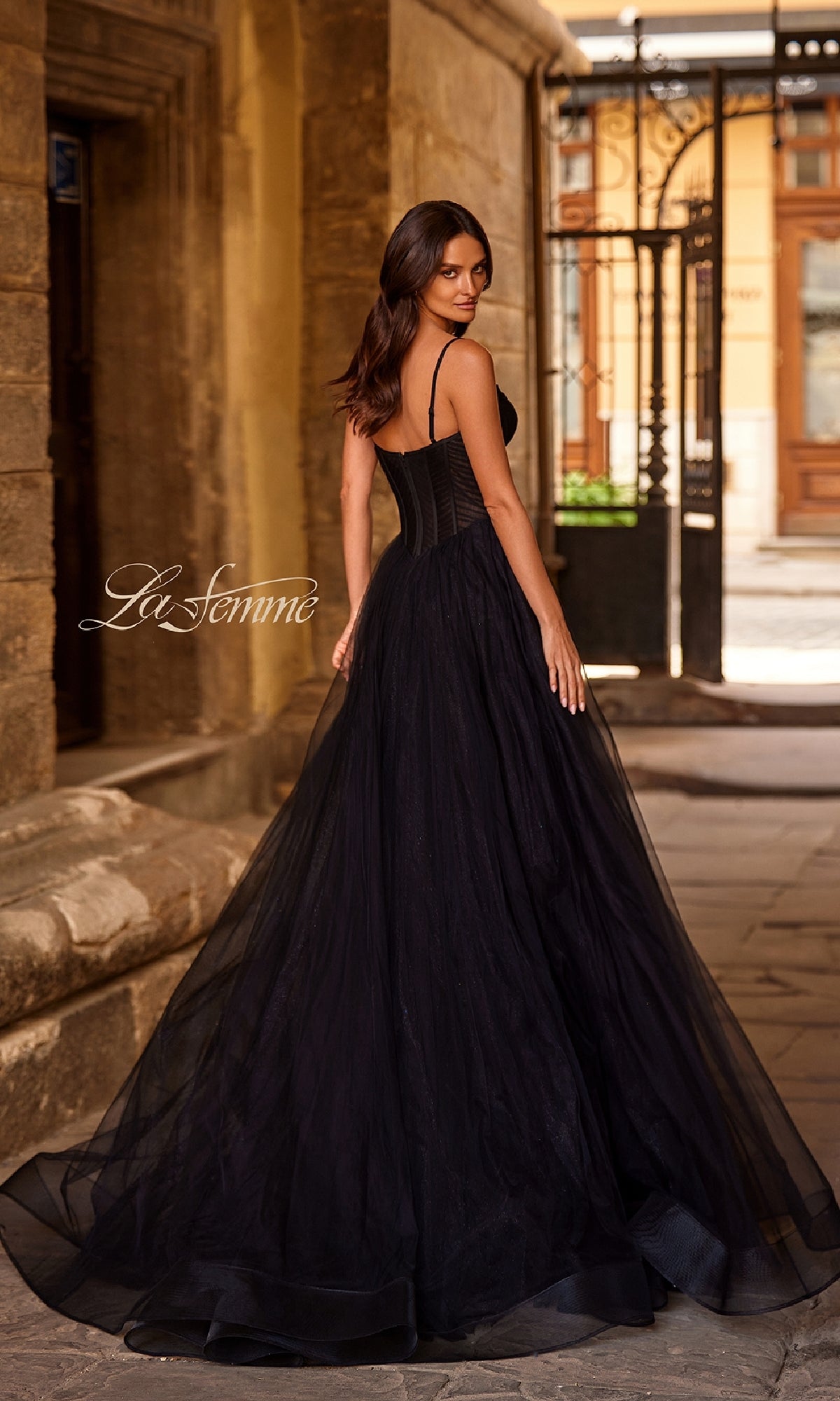 A woman in a Long Corset A-Line Prom Dress, La Femme 32680, with thin straps stands outdoors, looking over her shoulder in a historic stone courtyard.