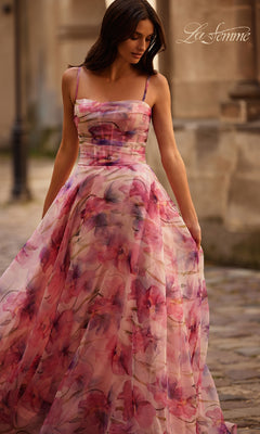 A woman stands outdoors on a cobblestone path wearing the La Femme 32686 pink floral-print corset prom dress.