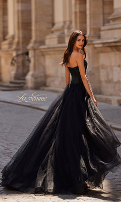 A woman in the La Femme 32690 Strapless Long Prom Ball Gown stands on a cobblestone street before a stone building, looking over her shoulder.