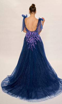 A woman in a dark blue Nina Canacci 3269 long formal dress featuring a low back, purple floral details, tulle overlay, embellished fitted bodice, and bow straps stands against a plain background.