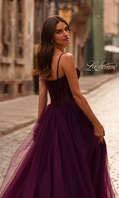 A woman in the La Femme 32700 Tulle Prom Ball Gown with Pockets, a flowing strapless deep purple dress, stands on a cobblestone street and looks back over her shoulder.