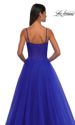 A woman faces away, wearing the La Femme 32700 Tulle Prom Ball Gown with Pockets in royal blue. This floor-length A-line dress features thin straps, a sheer corset-style bodice, and a white backdrop with the "La Femme" logo in the top right corner.
