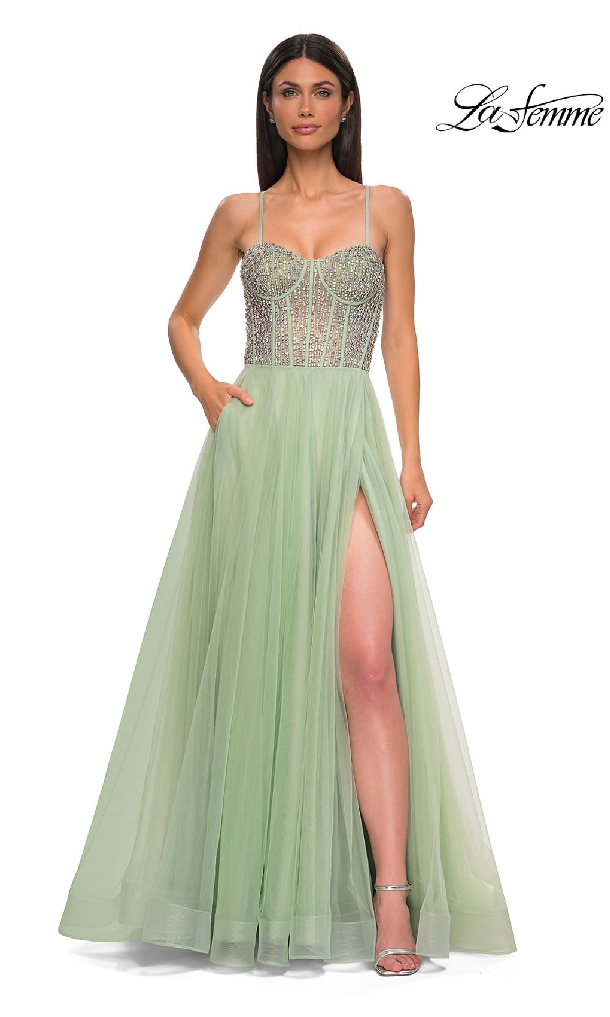 A woman models the La Femme 32703 Beaded-Corset Long Prom Dress—a light green evening gown with rhinestones, thin straps, and a thigh-high slit—while standing against a white background with the 