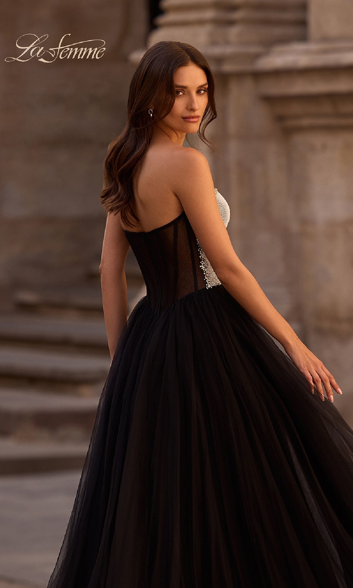A woman wearing the La Femme 32704 long prom dress stands outdoors, looking over her shoulder. The strapless gown features a flowing tulle skirt, with stone steps and a building in the background.