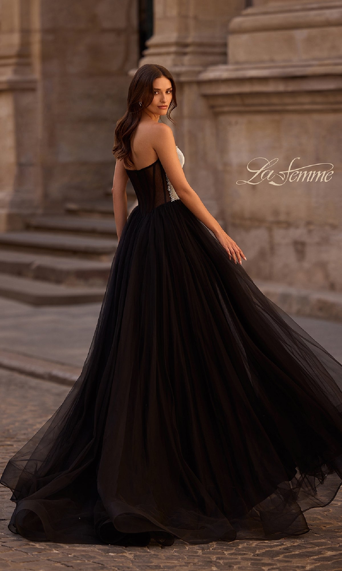 A woman in the La Femme 32704 long prom dress with a strapless bodice and flowing tulle skirt stands on a cobblestone street, glancing over her shoulder.