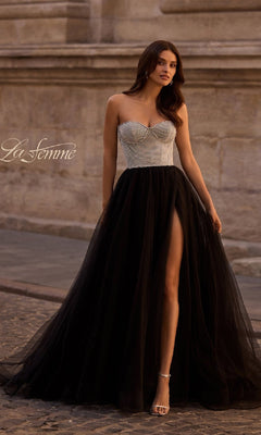 A woman stands on a cobblestone street in the La Femme 32704 long prom dress, featuring a strapless white beaded bodice and a black tulle skirt with a high slit.