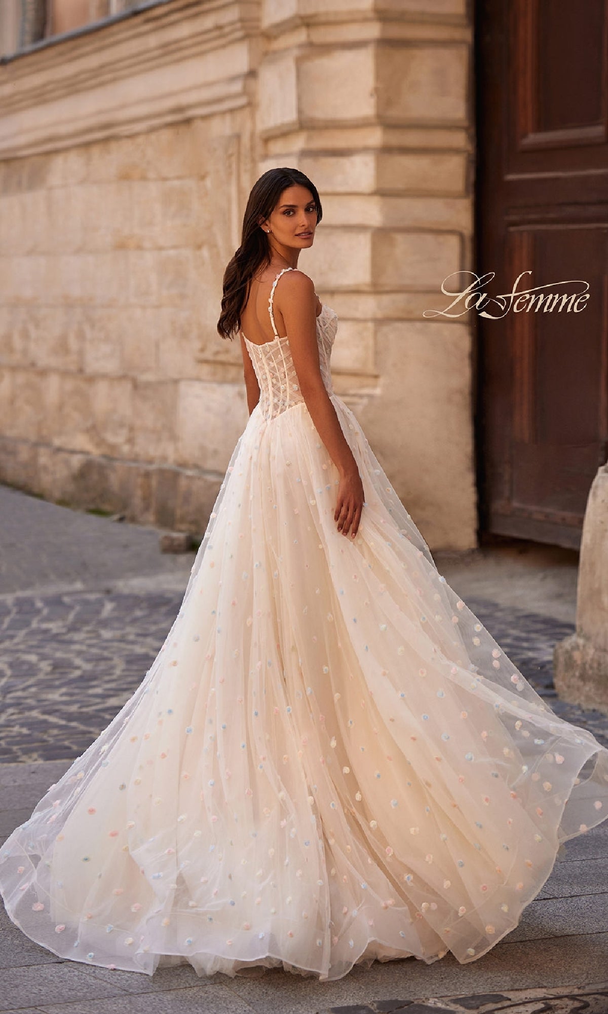 A woman in a Polka-Dot Long Prom Ball Gown: La Femme 32716 stands outdoors on a cobblestone path, looking over her shoulder.