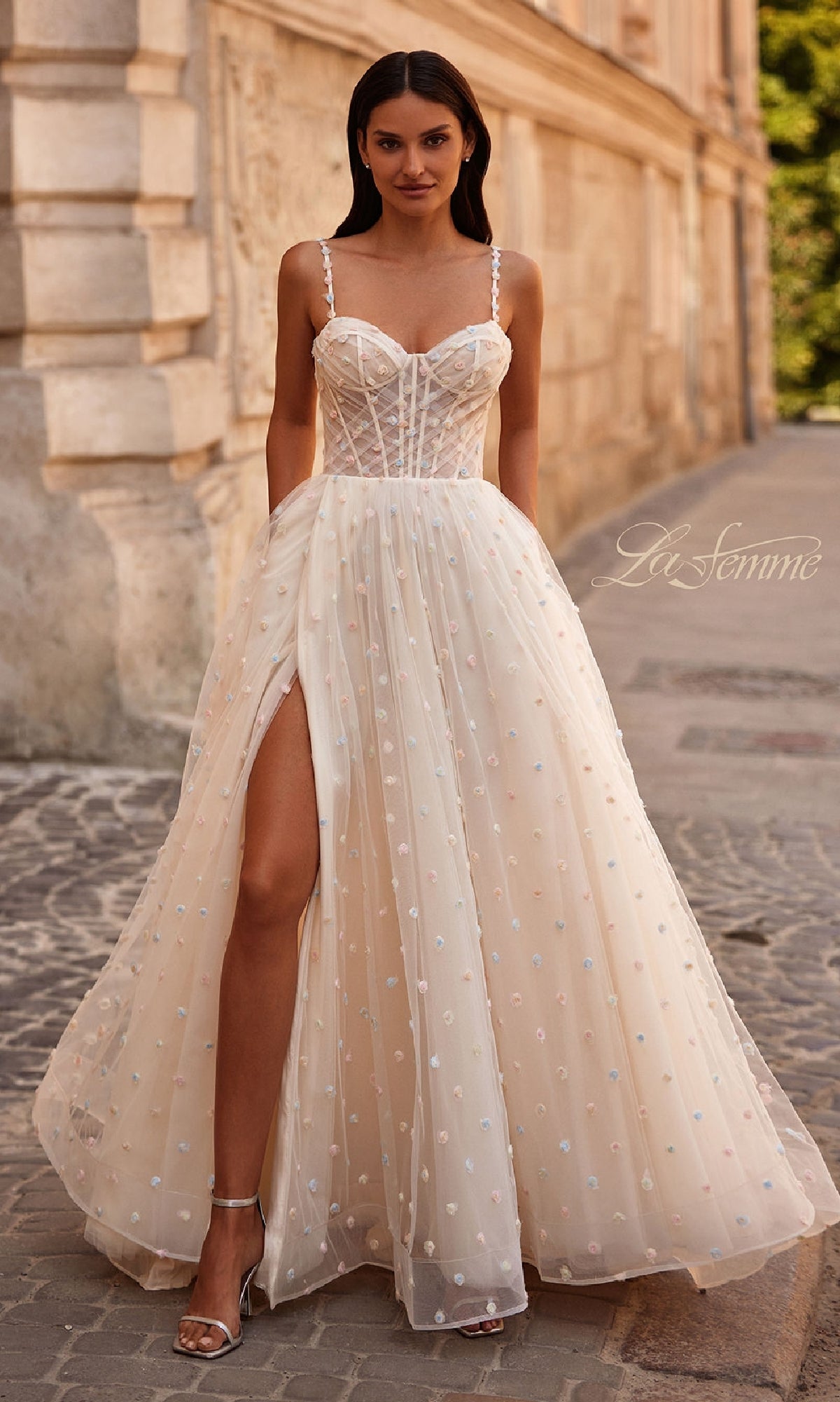 A woman in the Polka-Dot Long Prom Ball Gown: La Femme 32716 stands on a cobblestone path beside a stone building.