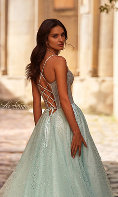 A woman stands outdoors on a cobblestone path, looking over her shoulder in the Lace-Up Beaded Long Prom Ball Gown: La Femme 32725, featuring a sparkly light blue design with a lace-up back.