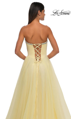 A woman in the pale yellow La Femme 32731 strapless long prom ball gown with a sweetheart neckline and lace-up back stands facing away, showcasing the floor-length, flowing fabric.