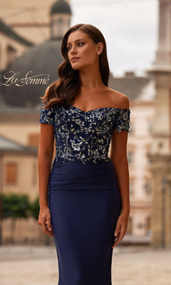 Outdoors in the city, a woman wears the Sequin-Bodice Long Navy Prom Dress (La Femme 32739)—an off-the-shoulder gown with a sparkling bodice that offers a perfect blend of sophistication and modern style.