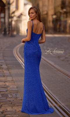 A woman wearing the Classic Long Beaded Prom Dress: La Femme 32753 stands on a cobblestone street, facing away. "La Femme" appears to the right, capturing the dress's elegance and sparkling blue allure.