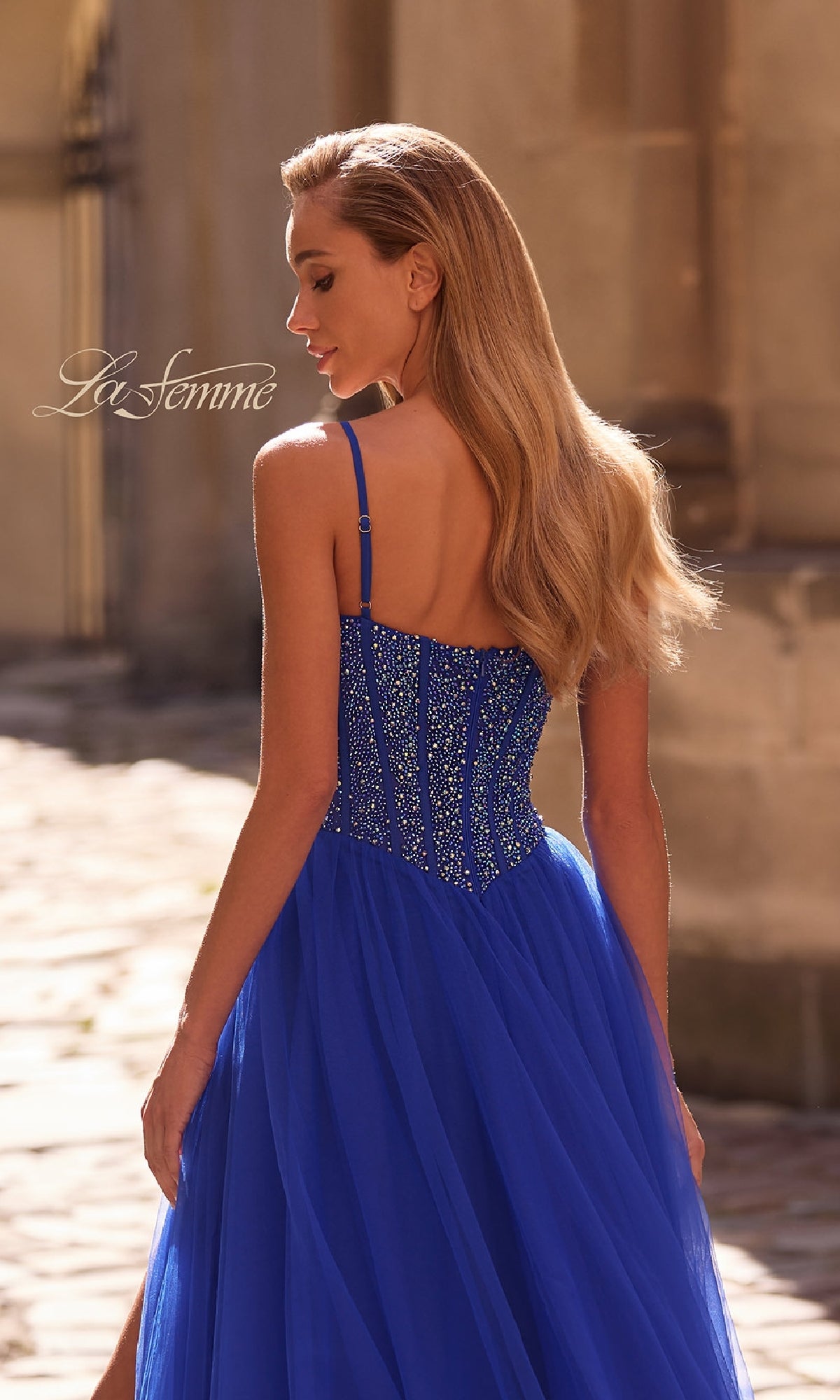 A woman in the La Femme 32754 Beaded-Bodice Long Prom Dress with a tulle skirt stands outdoors, back turned as sunlight highlights her long blonde hair.