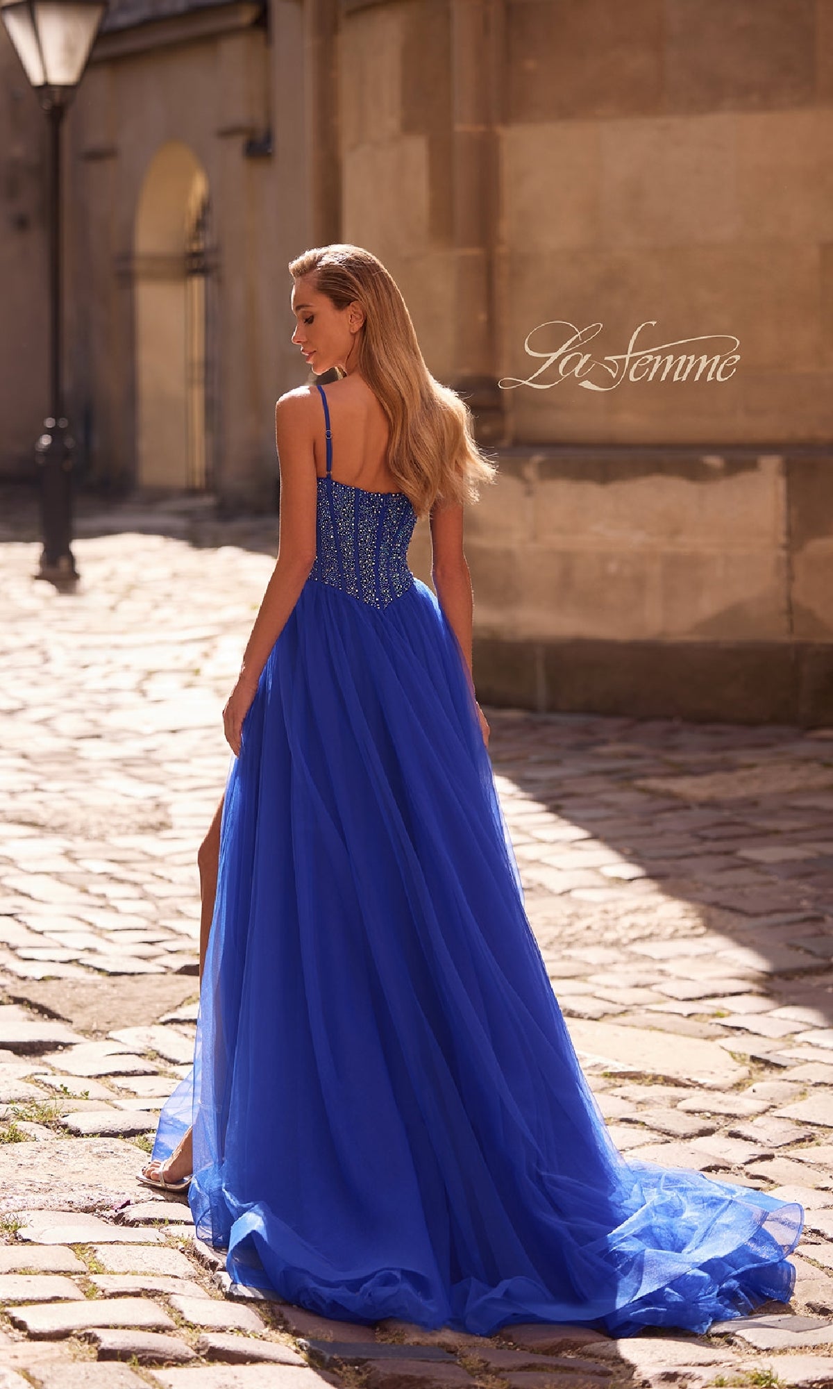 A woman in the La Femme 32754 Beaded-Bodice Long Prom Dress walks on a sunlit cobblestone street, her royal blue gown flowing elegantly as she faces away from the camera.