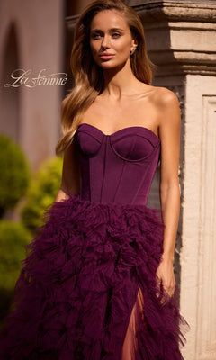 A woman stands outdoors near a stone column wearing the La Femme 32760, a strapless long ruffled prom dress in deep purple featuring a corset-style bodice and layered tulle A-line skirt.