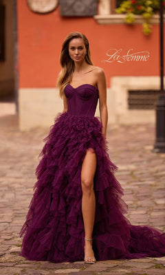 A woman stands outdoors on a cobblestone street, wearing the La Femme 32760 Strapless Long Ruffled Prom Dress with a thigh-high slit and corset-style bodice.