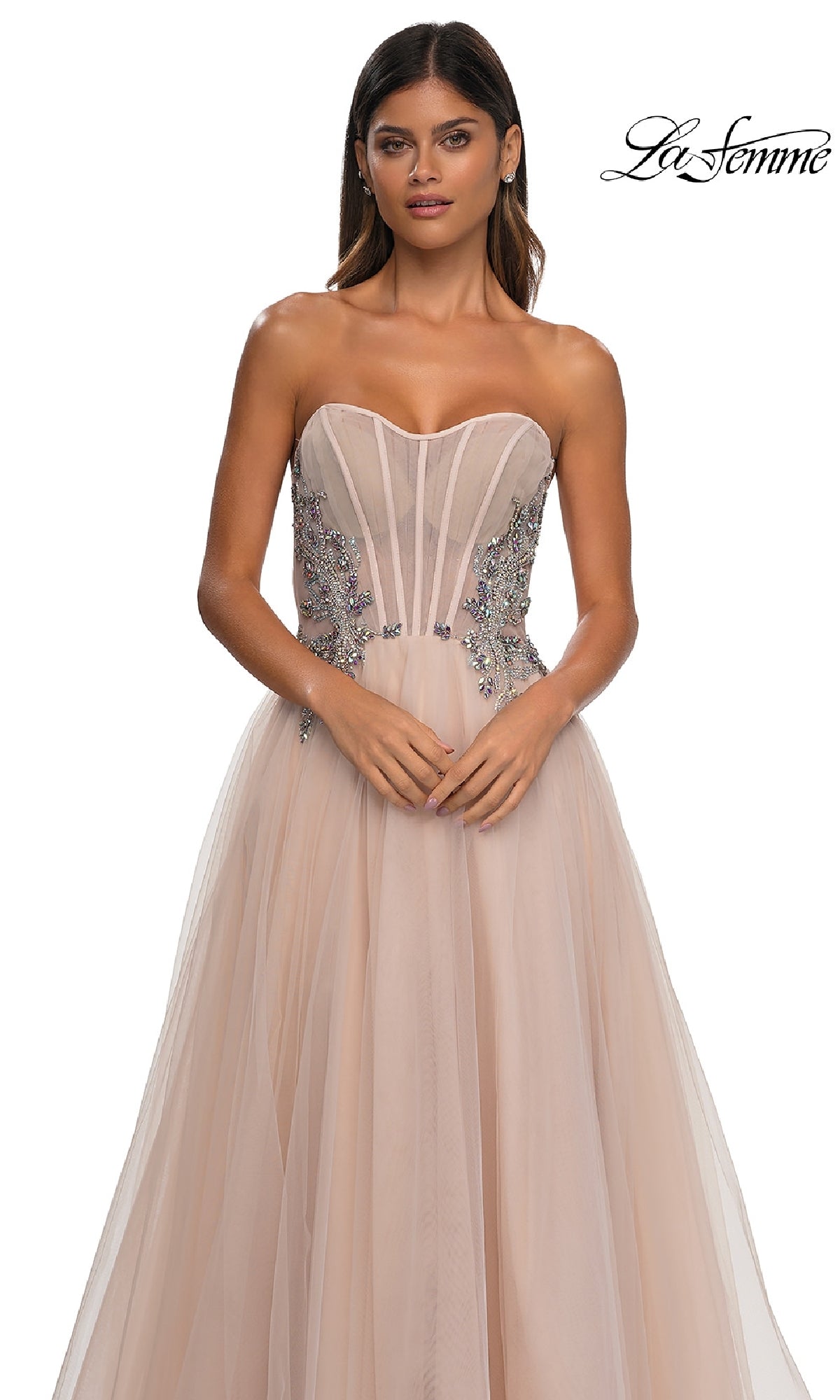 A woman wears the La Femme 32767 strapless long prom ball gown, featuring a blush hue, sheer beaded corset bodice, and floral embellishments, standing against a white background.