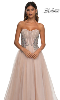 A woman wears the La Femme 32767 strapless long prom ball gown, featuring a blush hue, sheer beaded corset bodice, and floral embellishments, standing against a white background.