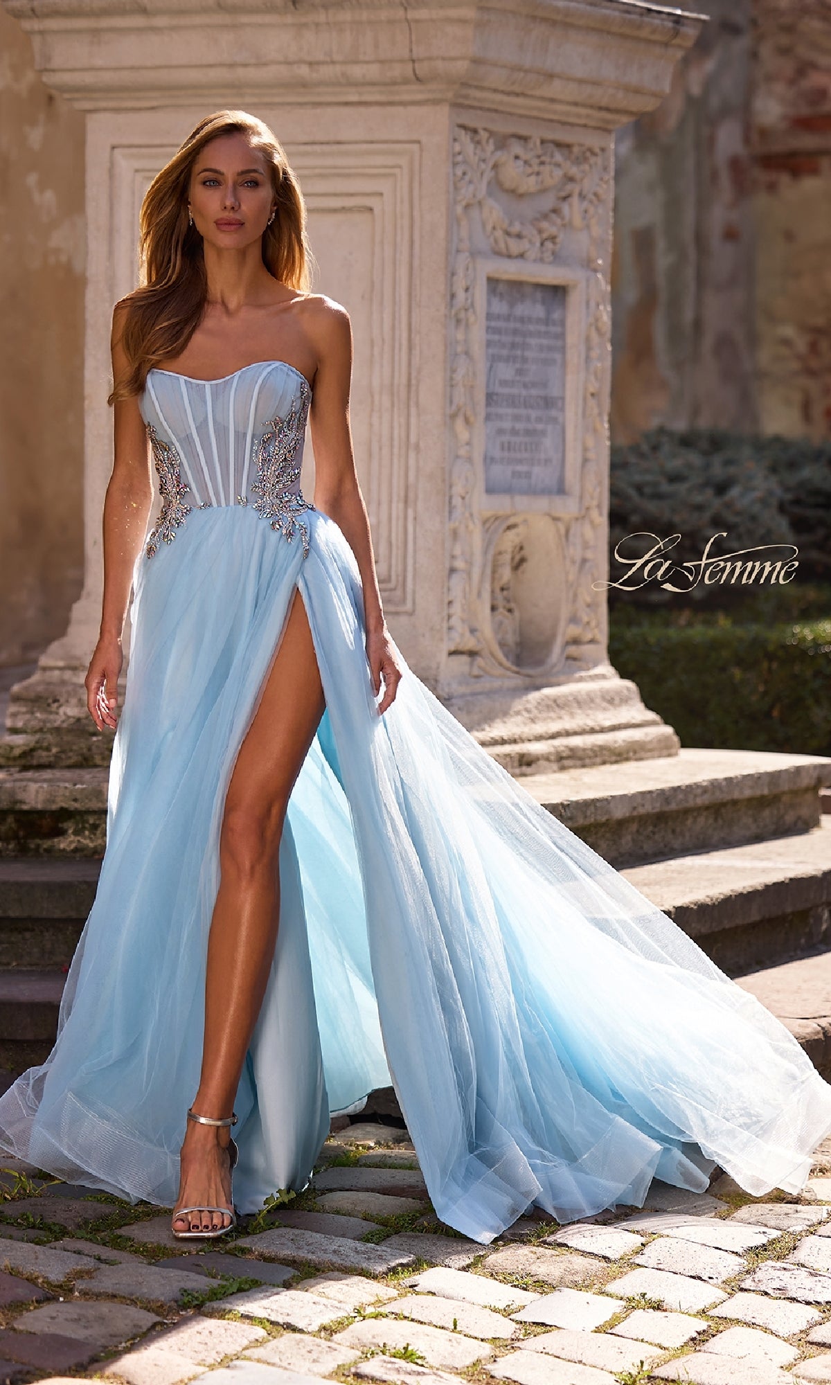 A woman wearing the La Femme 32767 strapless long prom ball gown with silver embellishments and a thigh-high slit stands outdoors near a stone monument.