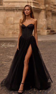A woman wearing the La Femme 32771 black strapless long prom dress with a sweetheart neckline and thigh-high slit walks on a cobblestone street in front of a stone building.