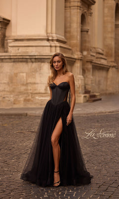 A woman wears the La Femme 32771 black strapless long prom dress with a sweetheart neckline, sheer skirt, and high slit as she stands on a cobblestone street before a historic stone building.