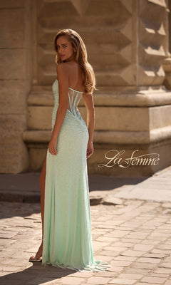A woman wears the La Femme 32774 strapless long beaded prom dress, featuring a side slit, while standing on a stone street and looking over her shoulder. The word "La Femme" appears in the image.
