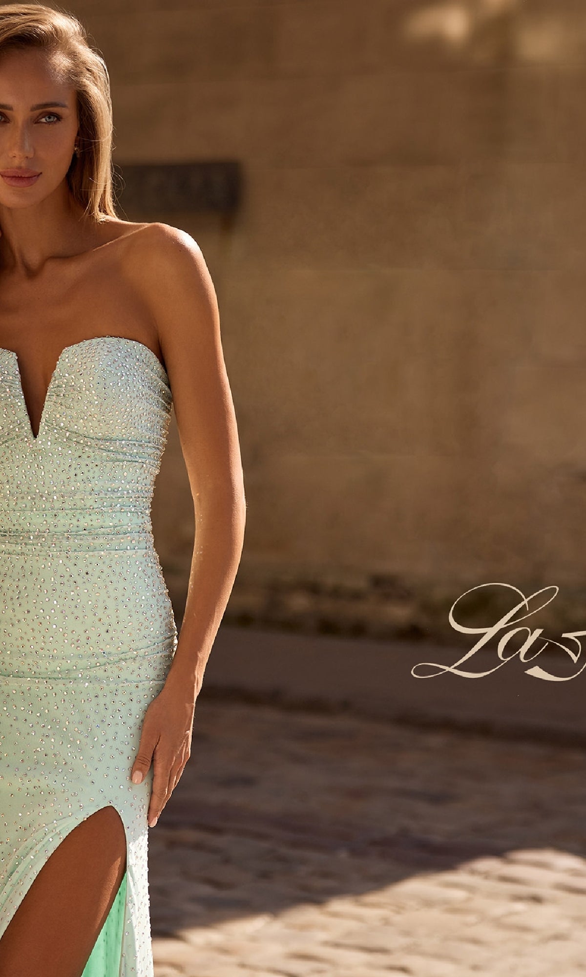 A woman in the Strapless Long Beaded Prom Dress La Femme 32774, featuring a light green hue, deep neckline, and thigh-high slit, stands outdoors on a cobblestone surface.