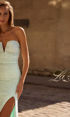 A woman in the Strapless Long Beaded Prom Dress La Femme 32774, featuring a light green hue, deep neckline, and thigh-high slit, stands outdoors on a cobblestone surface.