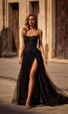 A woman stands outdoors on a sunlit stone street, her hair in loose waves, wearing the Long Black Glitter Prom Dress: La Femme 32781—a sleeveless gown with a high slit.